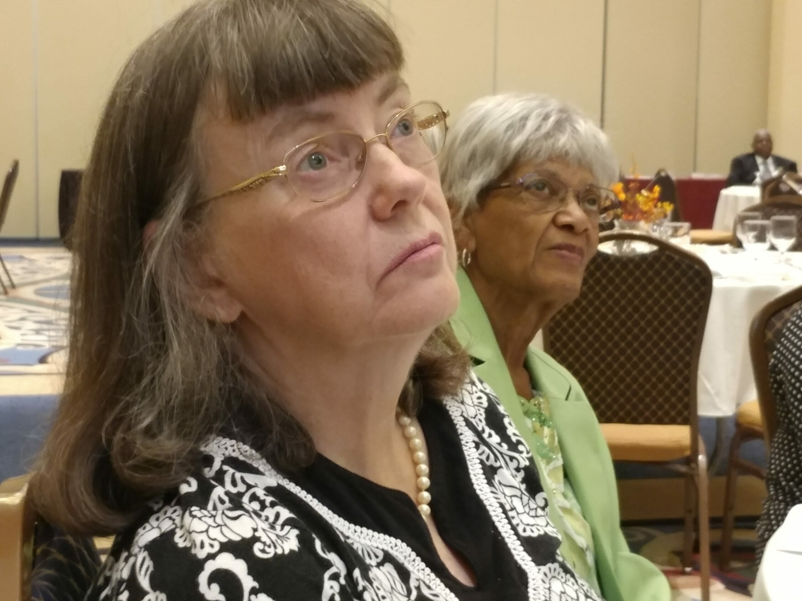 Two older women sitting at an event, paying attention to the event speaker.