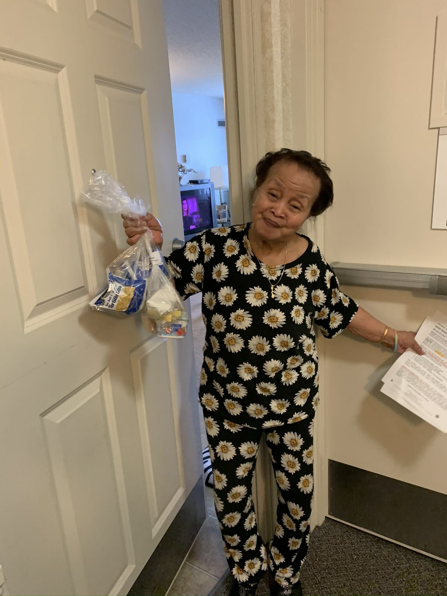 A short woman posing for the photo and holding bags in one hand and papers in the other hand.