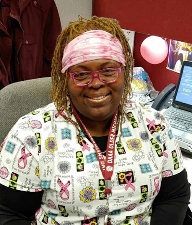 An African-American woman wearing scrubs is sitting at a desk, smiling at the camera.