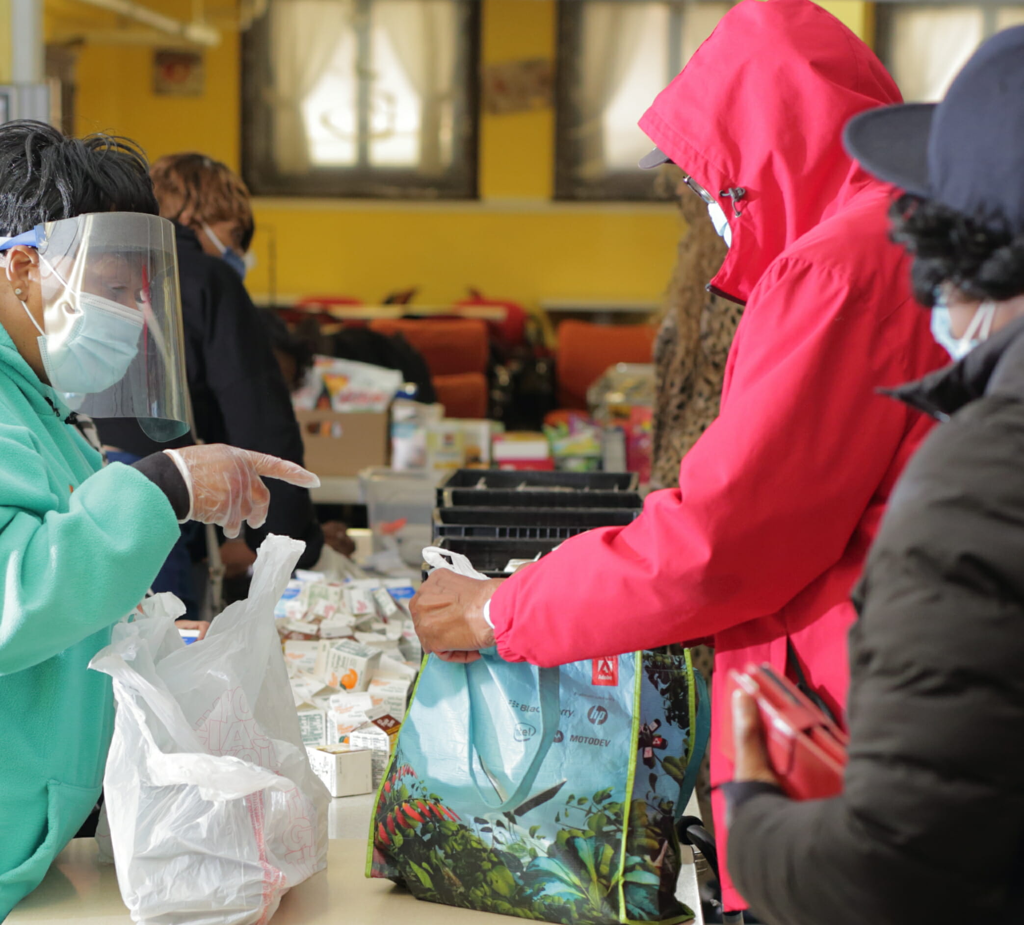 Individuals getting food to fill their reusable bags and DAAA volunteers helping to give out items | Image of a DAAA volunteer scooping food with their protective gloves and masks on | DAAA's Congregate Meal Program